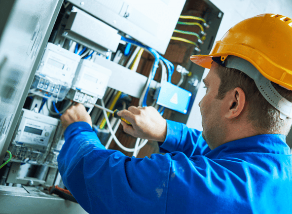 Ein Elektriker in Arbeitskleidung und mit gelbem Helm arbeitet am Elektroschaltkasten